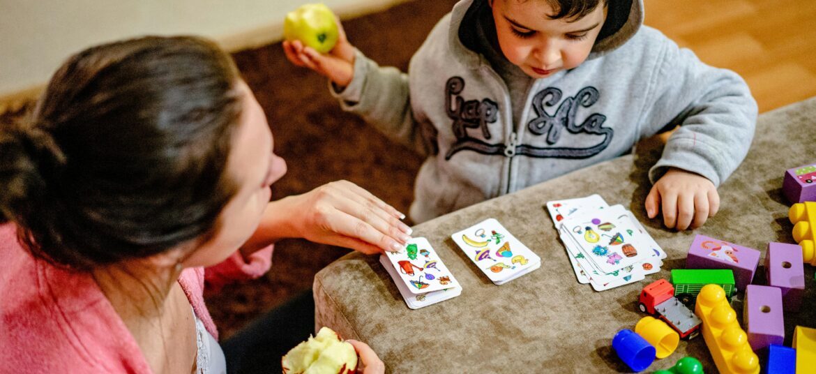Mother and child playing a card game surrounded by colorful toys indoors.