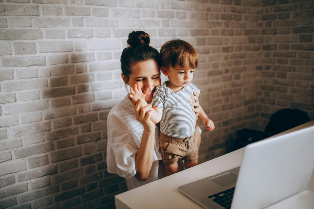 A mother waves at a laptop with her toddler, smiling warmly in their cozy home environment.